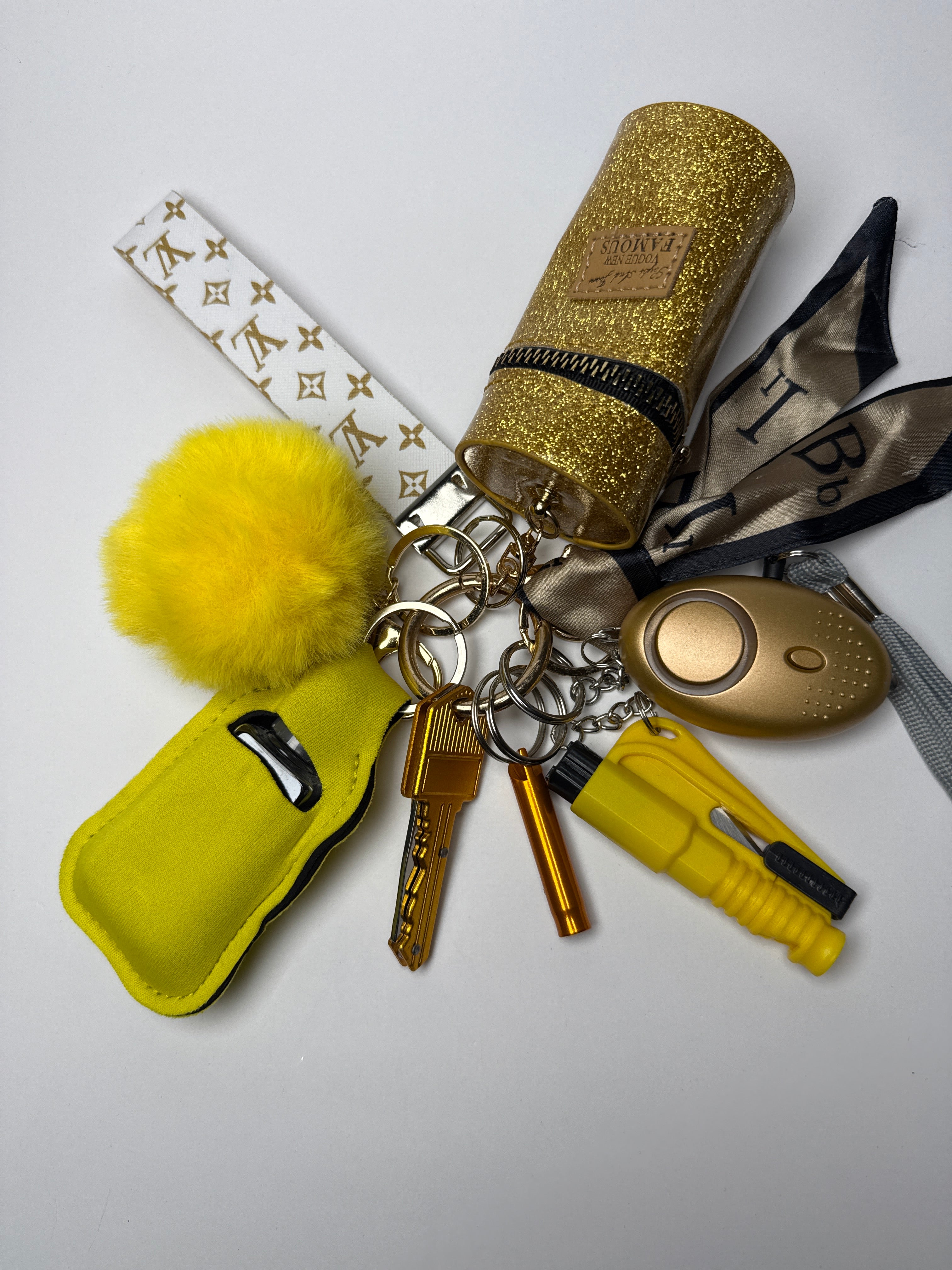 Gold wristlet keychain set with yellow pom charm, compact personal alarm, mini light, whistle, and glitter zip pouch on a white background