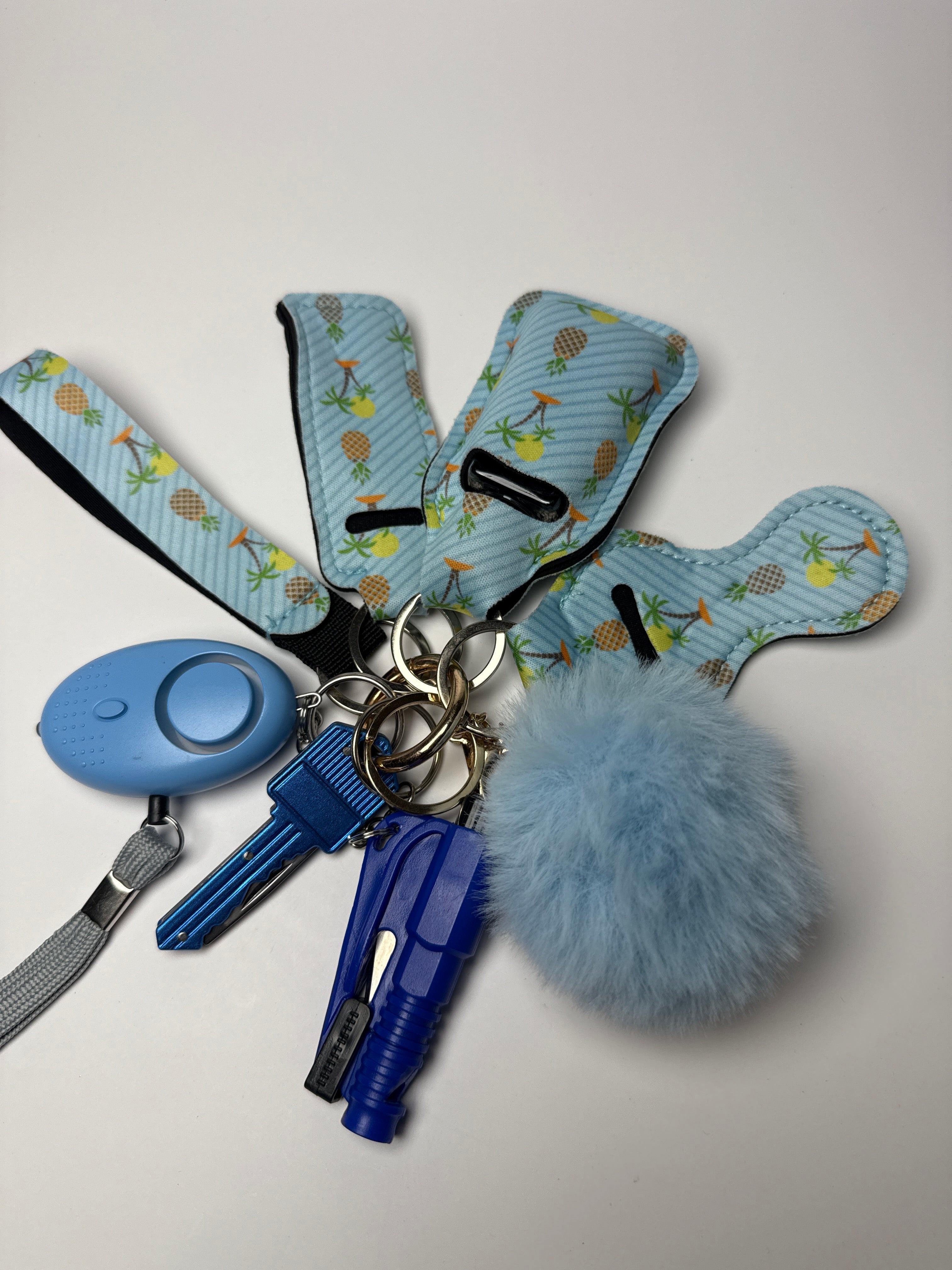 Pineapple print wristlet keychain set with light blue pom charm, compact personal alarm, and mini light on a white background.