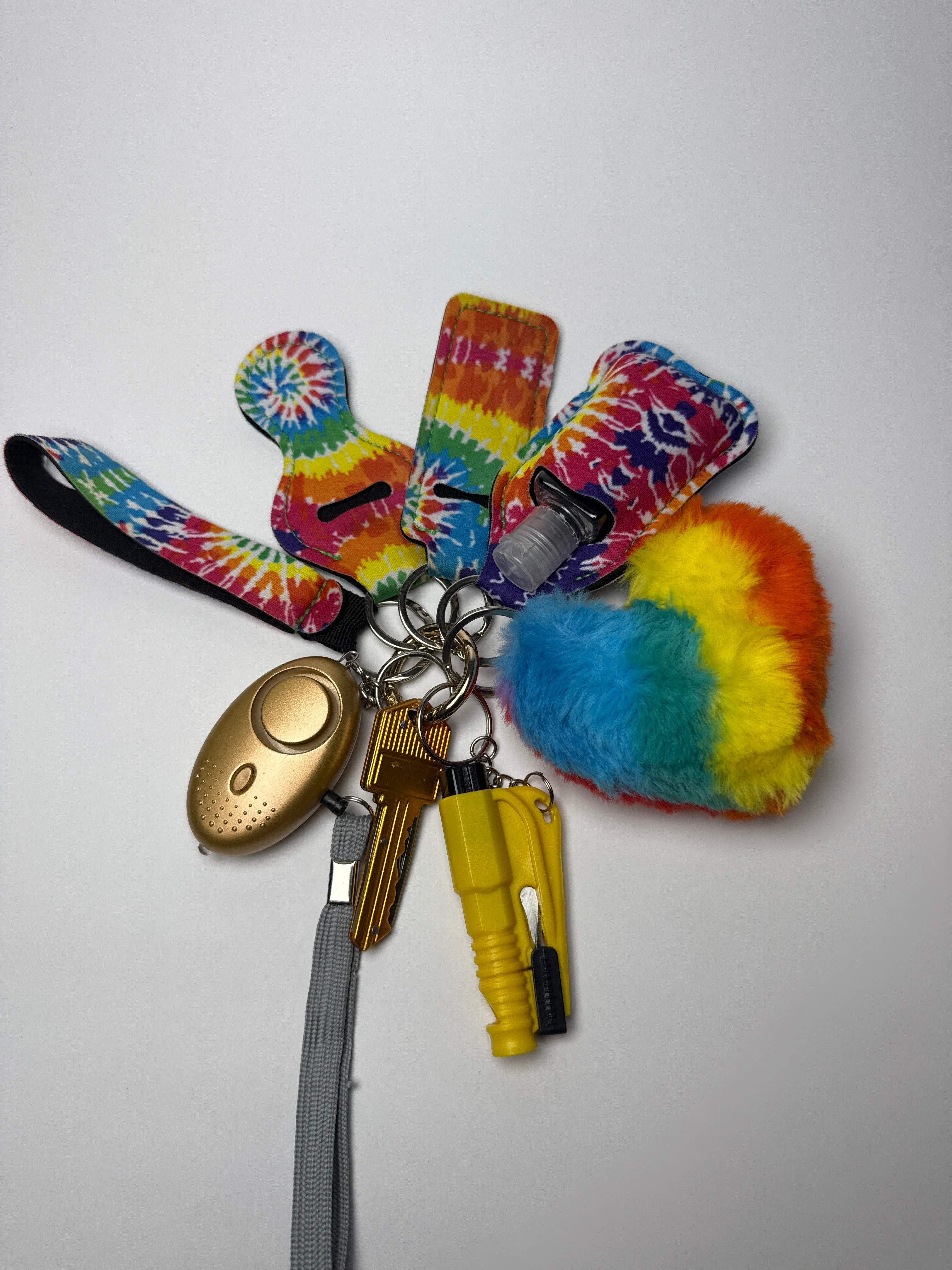 Multicolor tie-dye wristlet keychain set with rainbow pom charm, compact personal alarm, mini light, and clip-on bottle/holder on a white background.