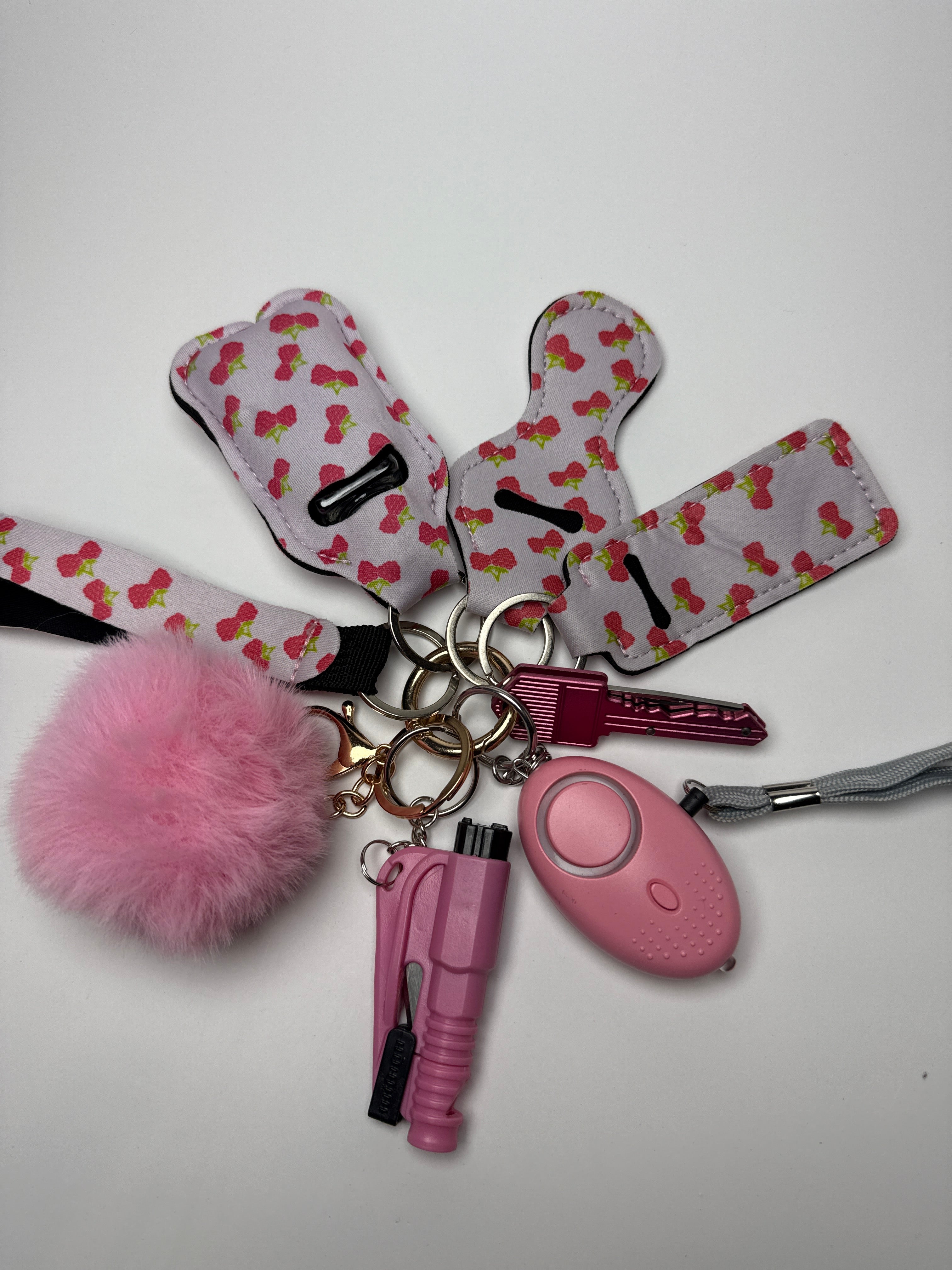 Cherry print wristlet keychain set with pink pom charm, compact personal alarm, and mini light on a white background.