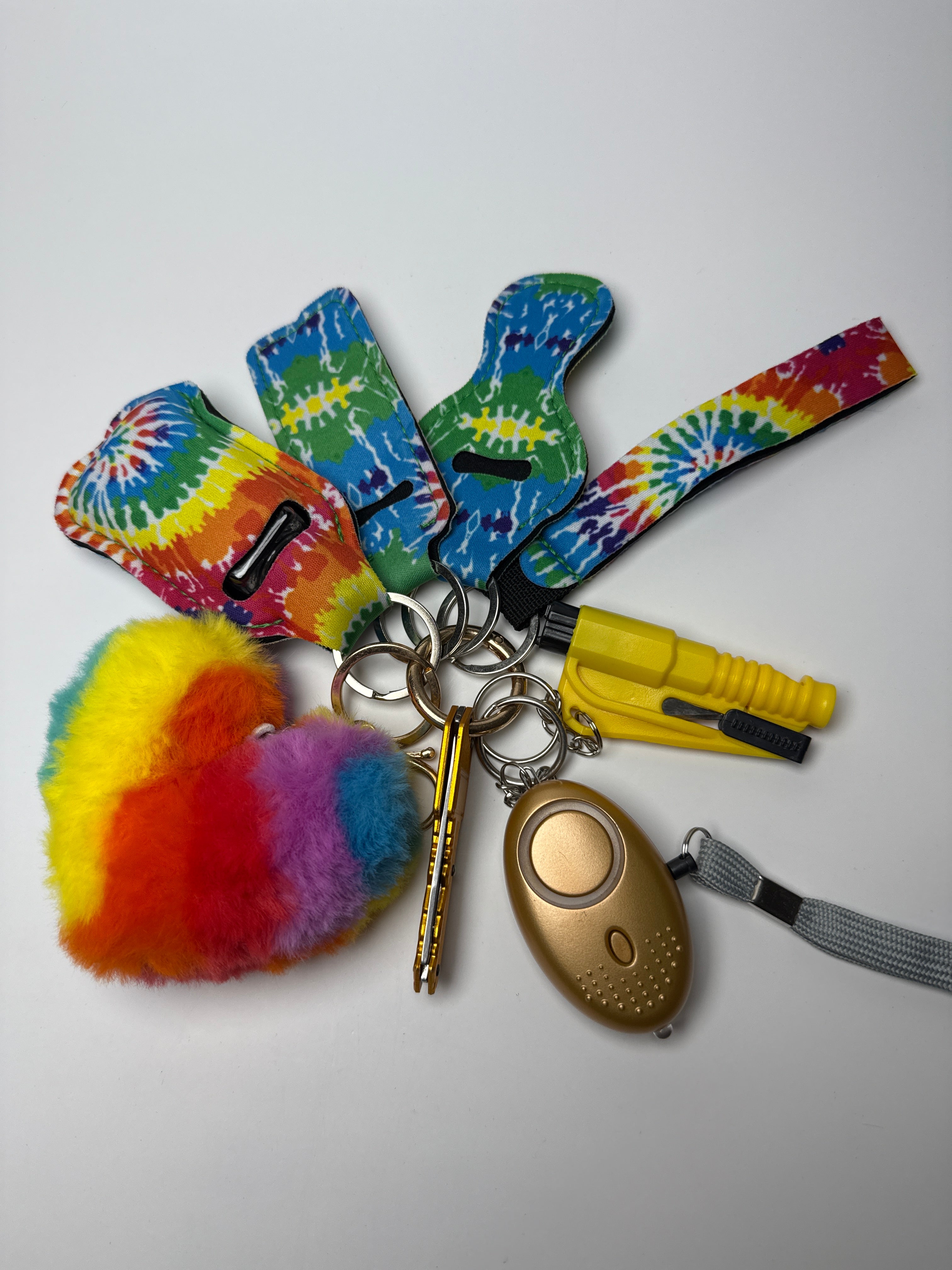 Multicolor tie-dye wristlet keychain set with rainbow pom charm, compact personal alarm, mini light, and clip-on bottle/holder on a white background.