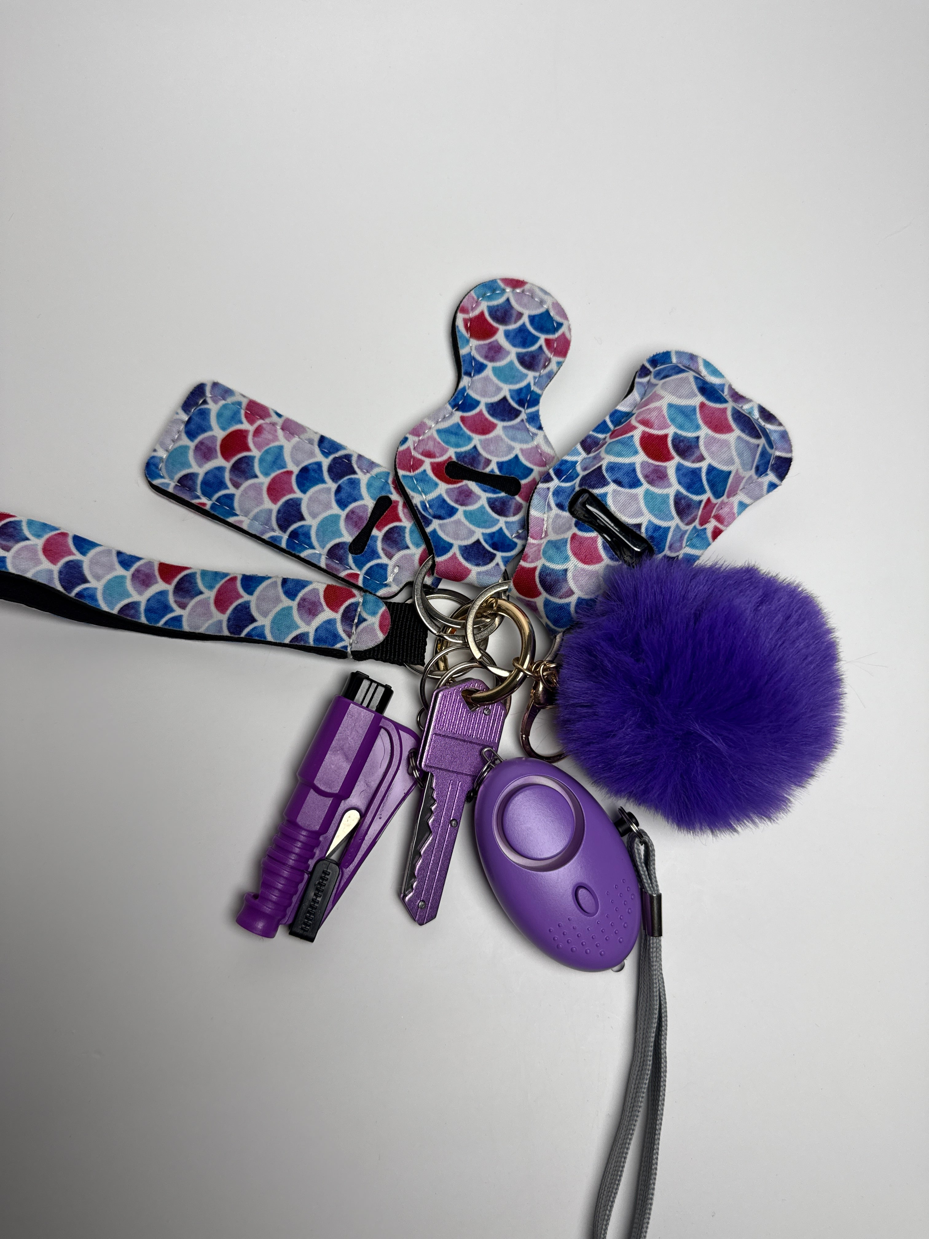 Mermaid scale wristlet keychain set with purple pom charm, compact personal alarm, and mini light on a white background.